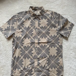 Beige mönstrad skjorta från East West - Kortärmad skjorta från East West i beige med stora svarta blommönster. Skjortan har klassisk krage, knäppning framtill och regular fit. Perfekt för dig som gillar unika prints och vill sticka ut lite extra.