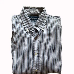 Randig skjorta från Ralph Lauren - Snygg skjorta från Ralph Lauren med klassiskt randigt mönster i vitt, blått och grått. Skjortan har lång ärm, knappar framtill och button-down krage. Broderad logga på bröstet ger en clean och stilren vibe. Perfekt för dig som gillar preppy stil.