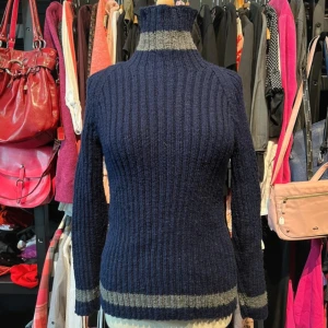 Marinblå handstickad polotröja med grå ränder - Snygg marinblå polotröja med ribbad struktur och gråa ränder vid hals, ärmslut och nederkant. Tröjan har lång ärm och hög krage som ger en cool och mysig vibe. Perfekt för dig som gillar klassisk stil med en twist. Tröjan är hand/hemstickad.