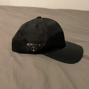 Svart keps från Prada i nylon - Snygg svart keps från Prada med klassisk böjd skärm och ikonisk metalltriangel på sidan. Tillverkad i glansigt nylonmaterial som ger en lyxig känsla. Perfekt accessoar för dig som gillar designer och vill sticka ut med en stilren look.