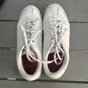 Vita cheer sneakers från Varsity - Säljer ett par helvita cheer sneakers från Varsity med snörning och låg profil. Skorna har rund tå, platt sula och är tillverkade i syntetmaterial. Perfekta för cheerleading eller träning. Diskret logga på sidan och sulan. Säljer då dem blivit för små.