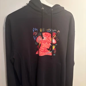 Svart Helmut Lang hoodie med tryck - Svart hoodie från Helmut Lang med färgstarkt grafiskt tryck på bröstet. Tröjan har huva med snörning och en klassisk känguruficka framtill. Perfekt för dig som gillar streetstyle och unika prints.