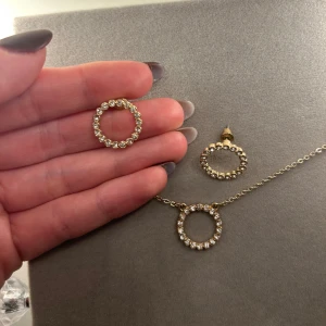 Guldigt smyckeset med cirklar och stenar - Säljer ett matchande set med halsband och örhängen i guldton. Både halsbandet och örhängena har en rund cirkelformad hängare dekorerad med gnistrande vita stenar. Kedjan är tunn och elegant, perfekt för att ge en glittrig touch till din stil.