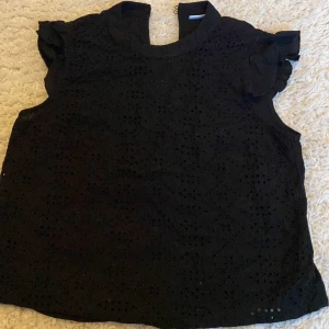 Svart blus med broderade detaljer - Svart blus från Jacqueline de Yong i storlek 36. Toppen har ärmlös design med volangdetaljer vid axlarna och ett broderat hålmönster framtill. Hög halsringning och liten öppning bak i nacken. Perfekt för dig som gillar stilrena men ändå unika plagg. 30kr med frakt 