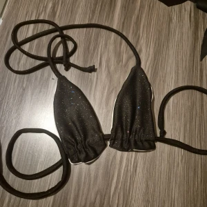 Svart-glittrig och vit bikini - Supersnygg bikini med trekantsmodell. Ena sidan är svart med glittriga detaljer, andra sidan är stilrent vit. Knytband i svart gör den enkel att justera. Perfekt för dig som vill kunna variera din look på stranden eller vid poolen. Skulle också fungera utmärkt för ex poledance. Helt oanvänd.