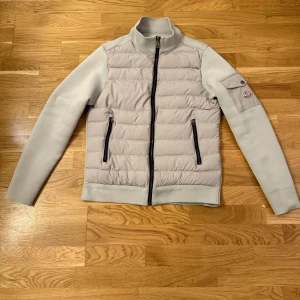 Ljusgrå pufferjacka från Moncler - Snygg ljusgrå pufferjacka från Moncler med quiltad front och släta ärmar. Jackan har svarta dragkedjor, hög krage och en ficka med Moncler-logga på ärmen. Materialmix med polyester och dun, perfekt för kyliga dagar.