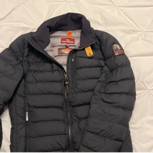 Svart lättviktsdunjacka Parajumpers S - Svart dunjacka från Parajumpers i lättviktsmodell med quiltad design och dragkedja framtill. Jackan har en snygg patch på ärmen och orange detalj vid kragen. Perfekt för dig som vill ha en stilren och funktionell jacka med grym passform. PytteLitet hål i höger ficka syns inte utifrån.