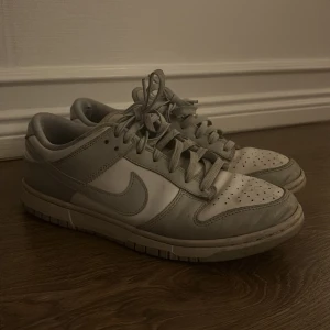 Nike Dunk Low i grå och vitt - Nike Dunk Low sneakers i en clean grå och vit färgkombination. Klassisk siluett med rund tå, perforerad tåbox och snörning. Ovandel i skinn och syntet, med den ikoniska swoosh-loggan på sidorna. Perfekta för dig som gillar streetstyle och tidlös design.