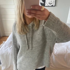 Grå stickad hoodie med huva - Mysig grå stickad hoodie med huva och snörning. Tröjan har en loose passform, långa ärmar och ribbade muddar vid ärmslut och nederkant. Perfekt för kyliga dagar och enkel att matcha med jeans eller leggings.