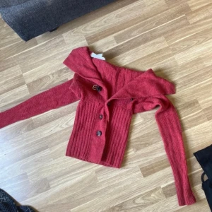 Röd stickad kofta med offshoulder - Säljer en röd stickad kofta med offshoulder-design och snyggt bälte med spänne vid kragen. Koftan har ribbade detaljer, stora knappar framtill och långa ärmar. Perfekt för dig som vill ha något unikt och trendigt i garderoben.