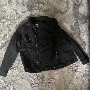 Svart jeansjacka från Zara - Svart jeansjacka från Zara med klassisk krage, knappar framtill och två bröstfickor med lock. Jackan har raka ärmar och en stilren look som passar perfekt till streetwear. Materialet är robust denim i svart färg.