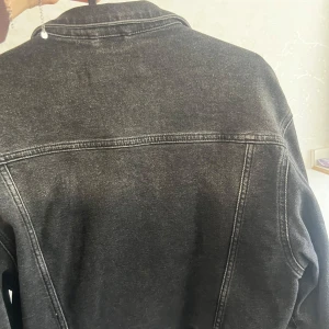Svart jeansjacka från Never Denim - Snygg svart jeansjacka från Never Denim i klassisk modell med dragkedja framtill och två bröstfickor med knappar. Jackan har en cool tvättad look och markerade sömmar som ger en trendig vibe. Perfekt för dig som gillar streetstyle.