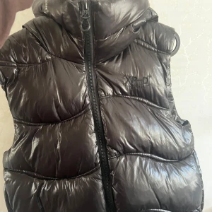 Svart dunväst från Superdry XP-D - Snygg svart dunväst från Superdry XP-D med glansig finish och vadderad design. Västen har huva, dragkedja framtill och diskret logga på bröstet. Perfekt för lager-på-lager och streetstyle.