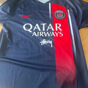 PSG x Stüssy x Nike fotbollströja - Snygg PSG fotbollströja i samarbete med Stüssy och Nike. Tröjan är marinblå med röd vertikal rand, PSG-logga på bröstet och Stüssy-tryck både fram och bak. Qatar Airways sponsortryck på framsidan. Tillverkad i lätt och ventilerande material. Storlek S men passar även M