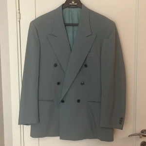 Blå dubbelknäppt kavaj från Corneliani - Snygg blå dubbelknäppt kavaj från Corneliani, tillverkad i Italien av extrafin merinoull (Super 100's). Klassisk modell med breda slag, fyra knappar framtill och diskreta fickor. Perfekt för dig som gillar stilrena och exklusiva plagg.