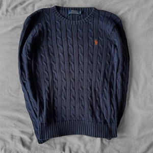 Mörkblå kabelstickad tröja Polo Ralph Lauren - Kabelstickad mörkblå tröja från Polo Ralph Lauren med klassisk orange broderad logga på bröstet. Tröjan har rund halsringning och är tillverkad i mjuk bomull. Perfekt för lager-på-lager och snygg till jeans eller kjol.