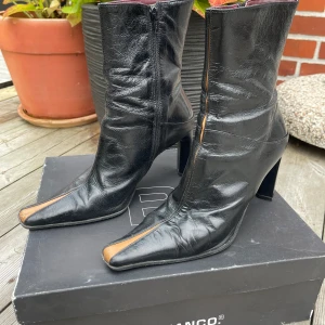 Svarta stövletter från Bianco med klack - Unika svarta stövletter i skinn från Bianco med spetsig tå och hög klack. Snygg detalj med brun rand längs tån och dragkedja på sidan. Insidan är fodrad i vinrött skinn. Köpta på 90-talet. 