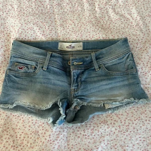 Blå jeansshorts från Hollister - Snygga ljusblå jeansshorts från Hollister med fransig kant och klassisk femficksdesign. Shortsen har låg midja, slitna detaljer och Hollisters logga på fickan. Perfekta för varma sommardagar och en avslappnad look.