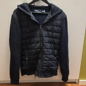 Mörkblå hoodie från Polo Ralph Lauren - Snygg mörkblå hoodie från Polo Ralph Lauren med quiltad framsida och dragkedja. Tröjan har huva med snörning, två fickor med knapp och mjuka ärmar. Perfekt för kyliga dagar och enkel att matcha med jeans eller joggers.