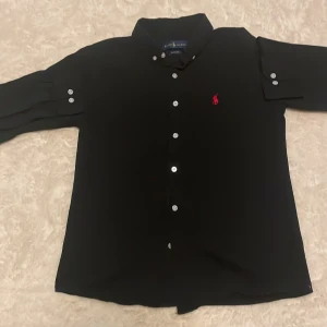 Svart skjorta från Ralph Lauren M - Svart skjorta från Ralph Lauren i klassisk passform med knappar framtill och vit knappdetalj på ärmarna. Ikonisk röd Polo-logga broderad på bröstet. Skjortan har krage och är tillverkad i bomull, perfekt för en clean och stilren look.