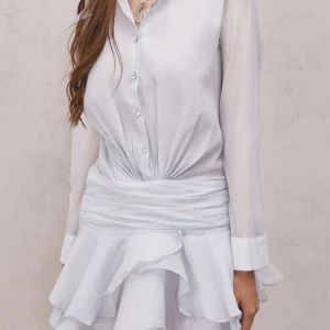 Vit omlottklänning med volang - Superfin vit kortklänning med långa ärmar, knappar framtill och markerad midja. Klänningen har volangig kjol och omlottliknande drapering i midjan. Tunt, lätt material som ger en luftig känsla och snygg passform.