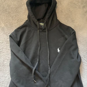 Svart hoodie från Polo Ralph Lauren - Svart hoodie från Polo Ralph Lauren med klassisk vit logga broderad på bröstet. Tröjan har huva med snörning och långa ärmar. Perfekt för en chill och stilren look.