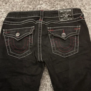 True Religion svarta jeans med kontrastsömmar - Slutsålda Svarta jeans från True Religion med vita och röda kontrastsömmar, ikoniska ficklock bak med stora knappar och broderade detaljer. Låg midja och bootcut-modell i klassisk jeanskvalitet. Snygga detaljer som ger en unik look. Inga tecken på andvändig, säljer för att jag knappt andvänder längre skorv om funderingar pris kan diskuteras vid snabb affär 