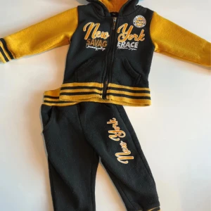 Svart och gul hoodie och byxor med New York-tryck - Svart och gul hoodie med huva och dragkedja framtill. Ärmar och nederkant har gula och svarta ränder. Stort New York-tryck på bröstet och matchande byxor med samma tryck och randiga muddar. Perfekt för en sportig och cool look.