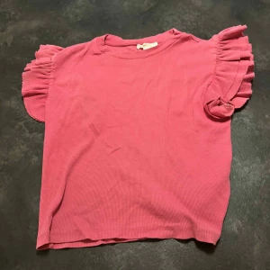 Rosa ribbad topp med volangärmar - Supersöt rosa topp med korta volangärmar och ribbad struktur. Toppen har rund halsringning och är tillverkad i mjukt material som känns skönt mot huden.