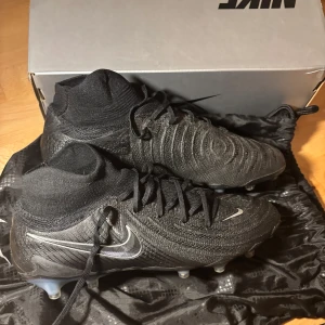 Nike svarta fotbollsskor med strumpkrage phantom luna II elite  - Säljer ett par svarta Nike fotbollsskor med strumpkrage och snörning. Skorna har en stickad ovandel och tydlig Nike-logga i silver på sidan. Sulan är utrustad med fasta dobbar för gräsplan och har blå detaljer undertill. Perfekta för dig som vill ha stilrena och moderna fotbollsskor.