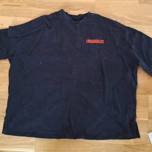 Svart t-shirt Los Angeles tryck Oversized XL  - Cool svart t-shirt med stor röd Los Angeles- och California-tryck på ryggen och ärmarna. Framtill finns en mindre Los Angeles-text i rött. T-shirten har rund hals och är kortärmad, tillverkad i mjuk bomull. Perfekt för dig som gillar streetstyle och bold prints.