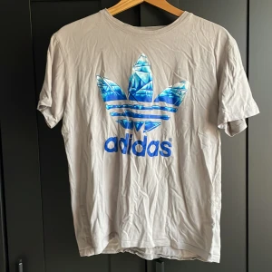 Grå Adidas t-shirt med blå logga - Snygg grå t-shirt från Adidas med stor blå glansig logga framtill. Klassisk rund hals och korta ärmar. Perfekt för dig som gillar sportig och avslappnad stil.