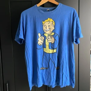 Blå Fallout t-shirt med Vault Boy - Säljer en blå t-shirt med stort tryck av Vault Boy från Fallout på framsidan. T-shirten har rund hals, korta ärmar och är gjord i mjuk bomull. Perfekt för dig som älskar gaming och vill sticka ut med en ikonisk design.