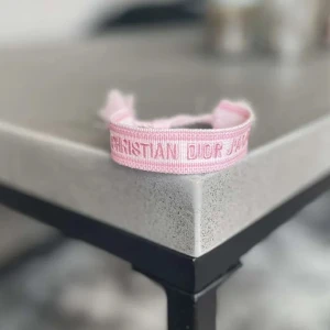 Rosa armband från Christian Dior - Snyggt rosa armband från Christian Dior med broderad logga i ljusrosa nyans. Armbandet är justerbart med knytband och har en trendig, ungdomlig stil som passar perfekt till din outfit.