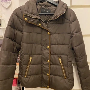 Brun pufferjacka från Vila - Snygg brun pufferjacka från Vila med guldiga dragkedjor och knappar. Jackan har quiltad design, hög krage och två fickor framtill med dragkedja. Perfekt för kyliga dagar och ger en cool vibe till din outfit.
