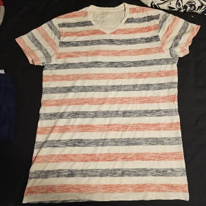 Randig t-shirt med v-ringning - Snygg Dressman t-shirt med v-ringad hals och korta ärmar. T-shirten har ett randigt mönster i vitt, rött och grått. Tillverkad i mjuk bomull, perfekt för en avslappnad stil. Passformen är normal och materialet känns lätt och skönt mot huden.