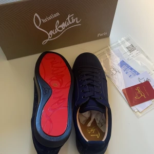 Christian Louboutin svarta sneakers - Svarta sneakers från Christian Louboutin med klassisk röd sula och broderad logga på hälen. Skorna har snörning och är tillverkade i mocka med rund tå och platt sula. Medföljer originalpåse, box och äkthetskort. Perfekt för dig som gillar exklusiva detaljer.