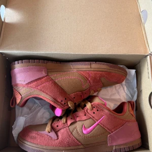 Nike Dunk Low i brunt och rosa - Nike Dunk Low sneakers i brunt och rödbrunt mocka med rosa detaljer och rosa Swoosh. Skorna har bruna snören, rund tå och platt sula. Extra skosnören i turkos och lila medföljer. Perfekta för dig som vill sticka ut med färg och stil.