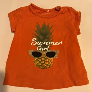 Orange t-shirt Summer Girl Name It - Snygg orange t-shirt från Name It med coolt ananastryck och texten 'Summer Girl' framtill. Ananasen har glittriga solglasögon för extra sommarkänsla. Gjord i mjuk ekologisk bomull och har korta ärmar. Perfekt för små trendsetters!