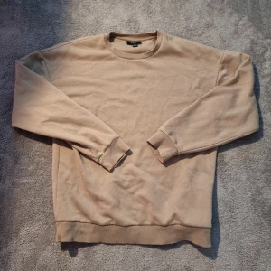 Beige sweatshirt från SMOG, stl S - Enkel och stilren beige sweatshirt från SMOG i storlek S. Tröjan har rund halsringning, ribbade muddar och är tillverkad i mjuk bomullsmix. Perfekt för en chill och avslappnad look.