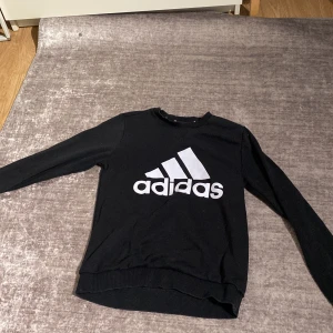 Svart Adidas sweatshirt med logga - Svart sweatshirt från Adidas med stor vit logga framtill. Tröjan har rund halsringning, långa ärmar och ribbade muddar vid ärmslut och nederkant. Perfekt för en sportig och avslappnad stil.