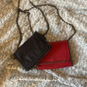 Stella McCartney axelväskor med kedja - Två snygga axelväskor från Stella McCartney, en i svart mocka och en i rött skinnimitation. Båda har coola kedjeremmar och dekorativa sömmar längs kanterna. Perfekta för att lyfta din outfit med en edgy vibe. !!50kr styck båda för 100kr!!