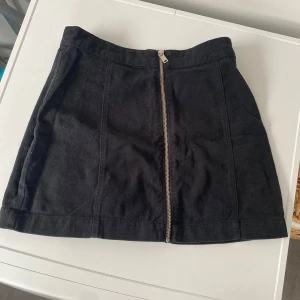 Svart kortkjol med dragkedja - Svart kortkjol i jeansmaterial med en snygg silverfärgad dragkedja framtill. Kjolen har en rak passform och är enkel men stilren, perfekt att matcha med en cool t-shirt eller stickad tröja. Midjehög modell som ger en trendig look.