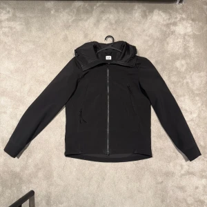 Svart softshelljacka från C.P. Company - Svart softshelljacka (C.P SHELL-R) från C.P. Company med huva och hel dragkedja framtill. Jackan har två snedställda fickor med dragkedja och är tillverkad i stretchigt syntetmaterial. Snygg, minimalistisk design som passar perfekt till streetwear. Äkta.