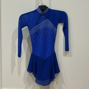 Blå glittrig konståkningsklänning - Superfin blå konståkningsklänning med långa ärmar, hög krage och öppen rygg. Klänningen är dekorerad med massor av glittrande stenar framtill och på kragen. Kjolen är kort och har ett flowigt fall. Perfekt för dig som vill sticka ut på isen!
