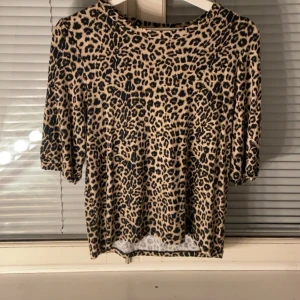 Leopardmönstrad topp med halvärm - Snygg topp med leopardmönster i beige och svart. Modellen har rund halsringning och halvärmar med lätt puff. Perfekt för dig som vill sticka ut med ett djurmönstrat plagg. Materialet känns mjukt och följsamt.