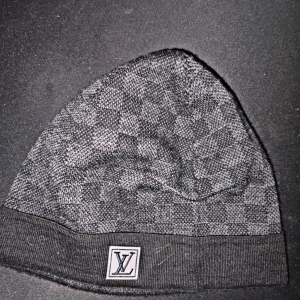 Louis Vuitton mössa - Snygg svart mössa från Louis Vuitton med diskret rutigt mönster och LV-logga framtill. Tillverkad i mjukt material som håller dig varm och stilren under kalla dagar. Perfekt accessoar för dig som gillar exklusiva detaljer.