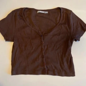 Brun croppad topp med knappar 157 - Brun croppad topp från 157 med korta ärmar och knappar hela vägen framtill. Toppen är ribbad och har en djup rundad ringning. Perfekt för dig som gillar en enkel men trendig stil.