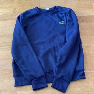 Blå sweatshirt från Lacoste - En klassisk marinblå sweatshirt från Lacoste med den ikoniska krokodilloggan broderad på bröstet. Tröjan har rund hals, långa ärmar och ribbade muddar vid ärmslut och nederkant. Perfekt för en chill och stilren look.