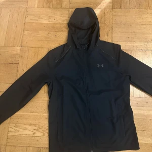 Under Armour jacka  - Svart vindjacka från Under Armour med huva och hel dragkedja framtill. Jackan har diskret logga på bröstet och stor UNDER ARMOUR-text på ryggen. Tillverkad i lätt och smidigt syntetmaterial, perfekt för aktiva dagar. Snygg och sportig look med lång ärm och normal passform.